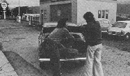 Black and white photo of two people putting spare tyre in car boot.