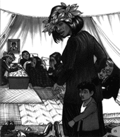 Body lies in state in a tent, surrounded by woven mats and mourners. A small child stands next to a woman with leaves in her hair in the foreground.